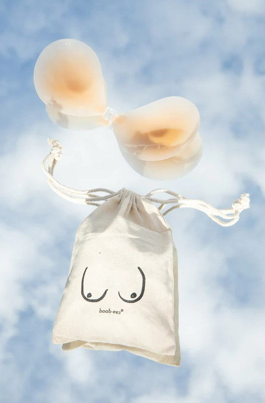 Product shot of Boob-eez Light Tint Silicone Stick On Bra with logo printed canvas bag on cloud background.