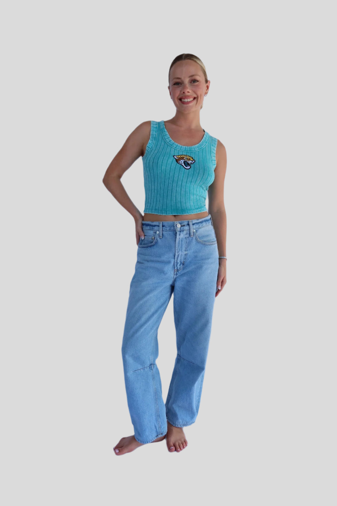 Woman wearing a turquoise tank top with a logo and blue jeans on a light background