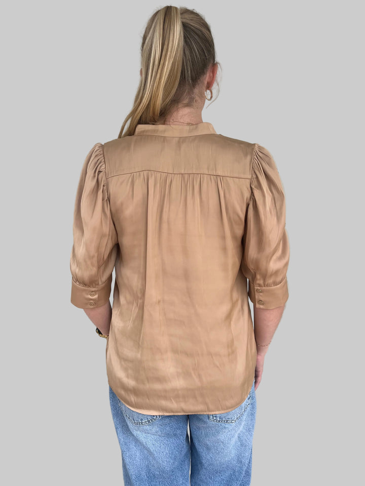 Woman wearing a bronze satin top and blue jeans against a light gray wall.