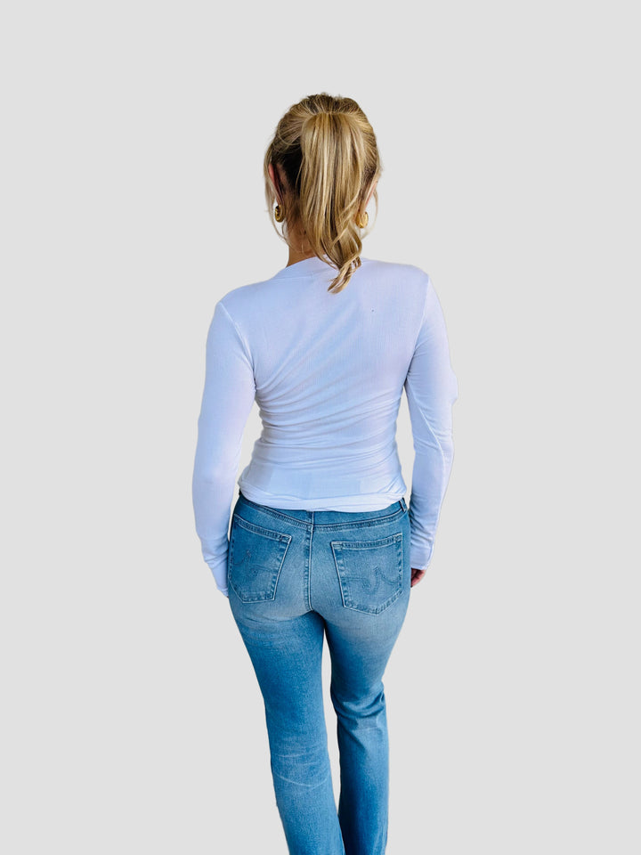 Person wearing a white long-sleeve top and blue jeans standing in front of a beige wall.