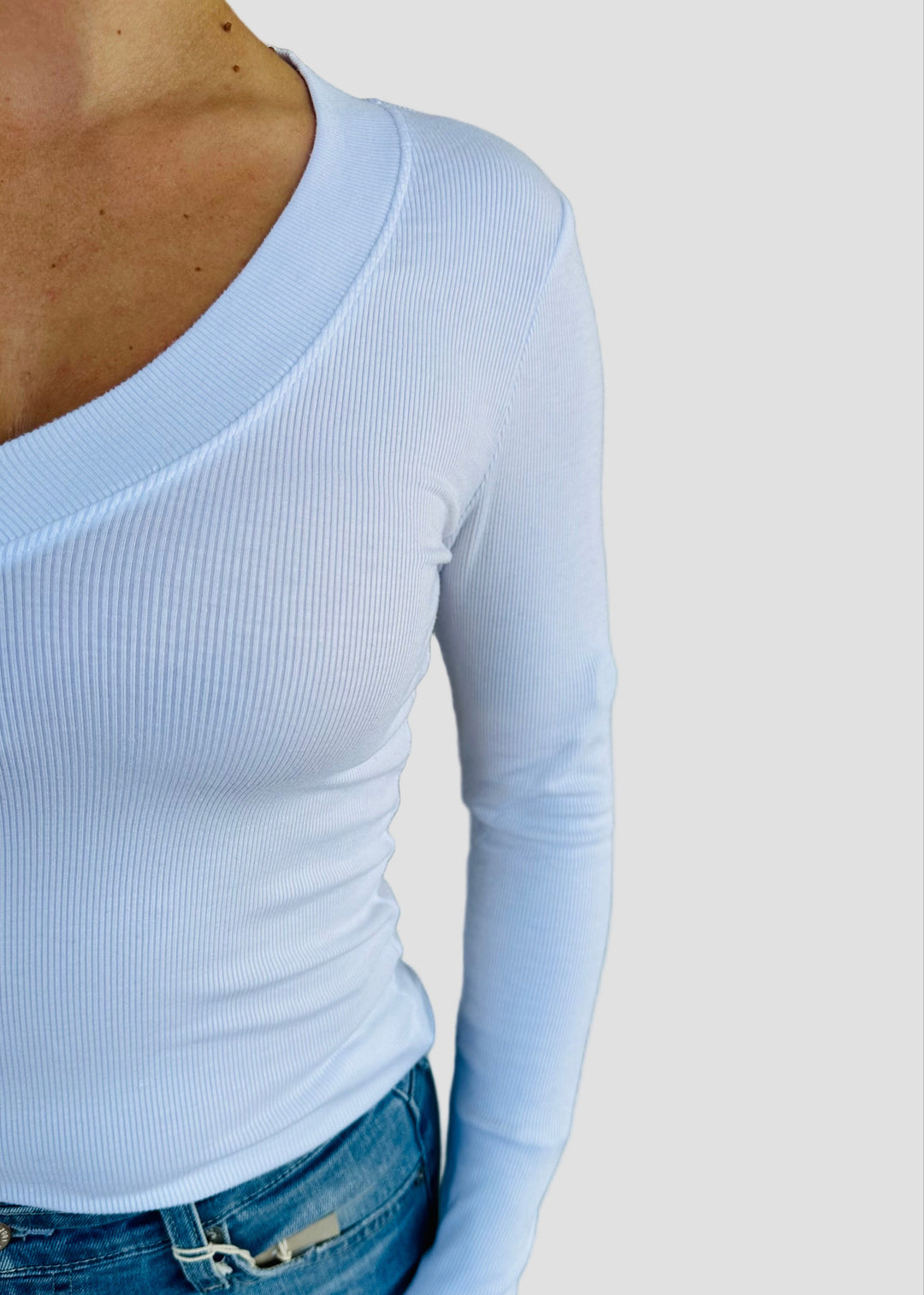Person wearing a white long-sleeve top and blue jeans against a light green wall.