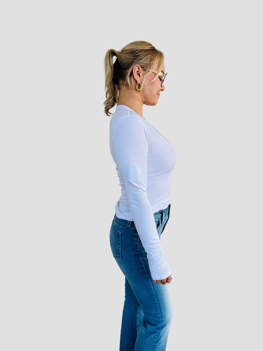 Woman wearing a white long-sleeve shirt and blue jeans standing against a beige wall.