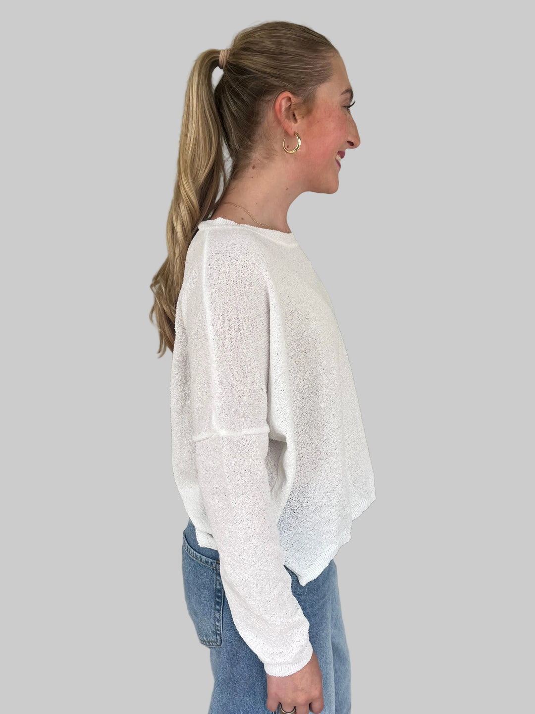 Person wearing a white long-sleeve Knit Top and blue jeans against a light gray wall.
