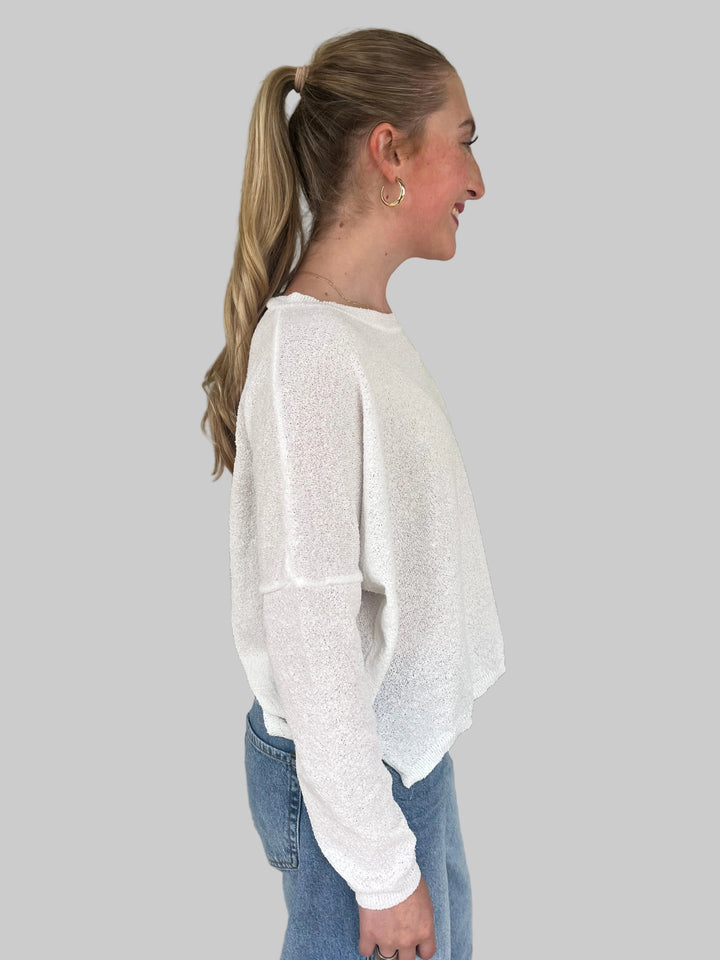 Person wearing a white long-sleeve Knit Top and blue jeans against a light gray wall.